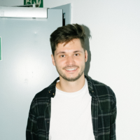 Max Sommer, a senior full-stack software developer, smiling and wearing a white t-shirt with a black checkered shirt, standing against a light grey background with a door slightly open behind him